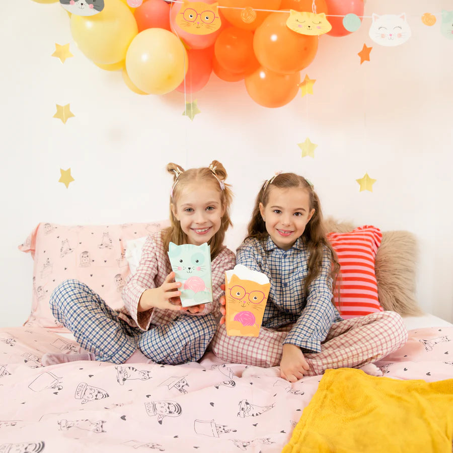 Two girls in pajamas holding popcorn paper boxes decorated with cats, sitting on a colorful bed with balloons in the background.
