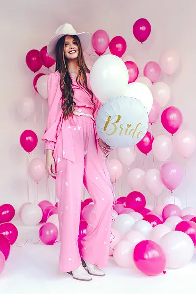 Happy bride holding round foil balloon amid pink and white balloons, dressed in pink outfit with a fashionable hat.