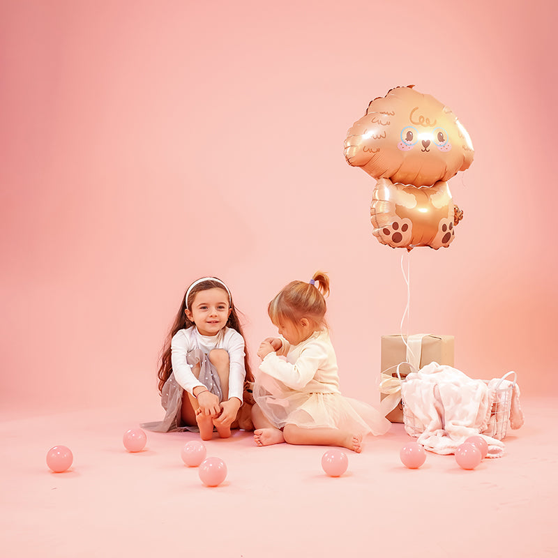 Two children sitting on a pink background with a foil balloon dog poodle, surrounded by decorative pink balls.