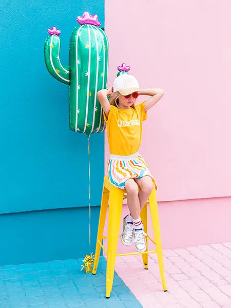 Child sitting on a yellow stool next to a bright green foil cactus balloon, vibrant pink and blue background.