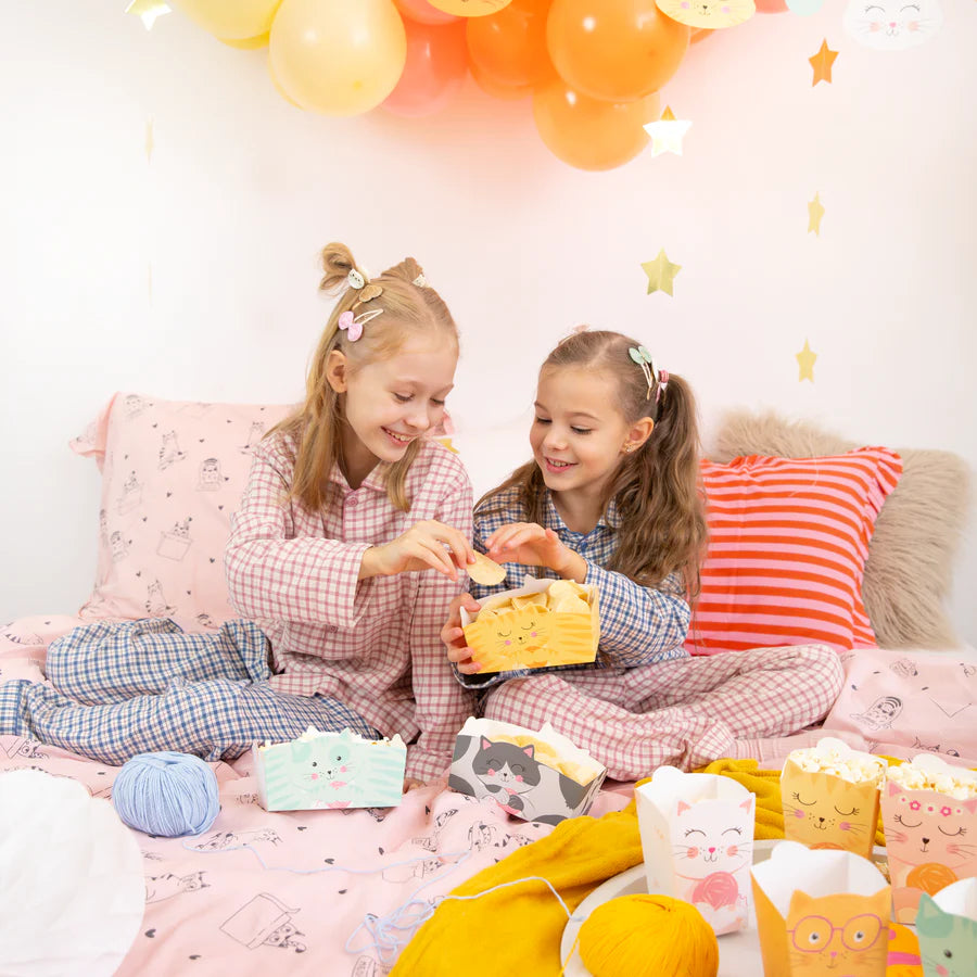 Two girls enjoying a fun moment with cat-themed chip boxes on a cozy bed, surrounded by playful decor.