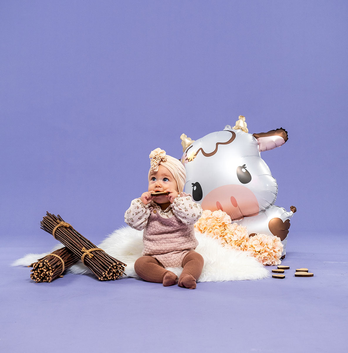 Adorable baby with a pink headband sitting next to a large foil cow balloon, surrounded by decorative sticks and flowers.