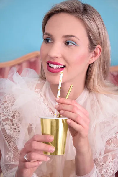 Model smiling and sipping from a gold cup with white paper straw featuring gold hearts.